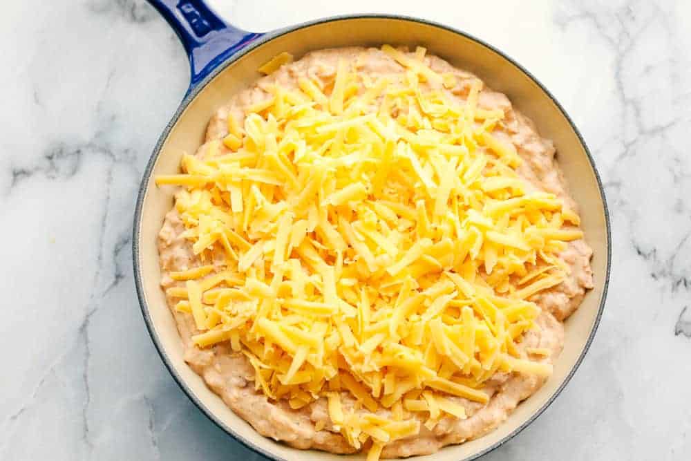 Process shot of bean dip in a blue skillet with cheese on top before baking.