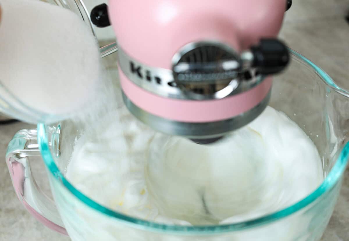 Angle shot of sugar being poured into stand mixer bowl full of whipped egg whites. 