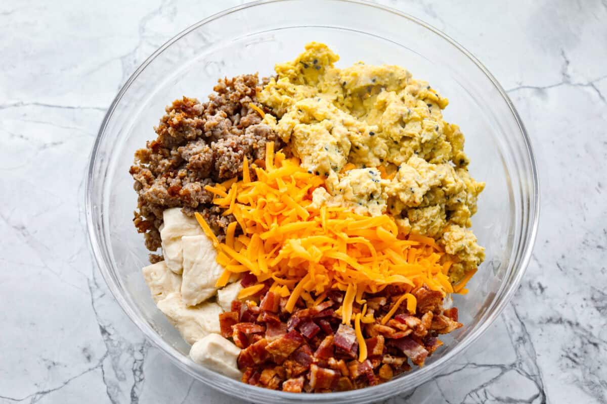 Overhead shot of sausage, bacon, eggs, cheese and biscuits separated in a bowl. 