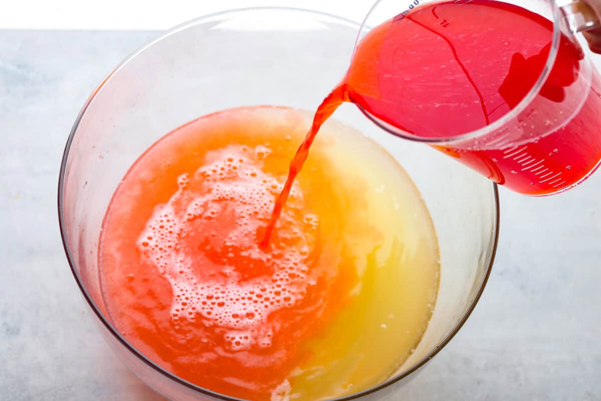 This is an overhead shot of a large bowl that has Sprite and pineapple juice in it. There is also a large measuring cup tipping its contents of Hawaiian Punch into the large bowl.