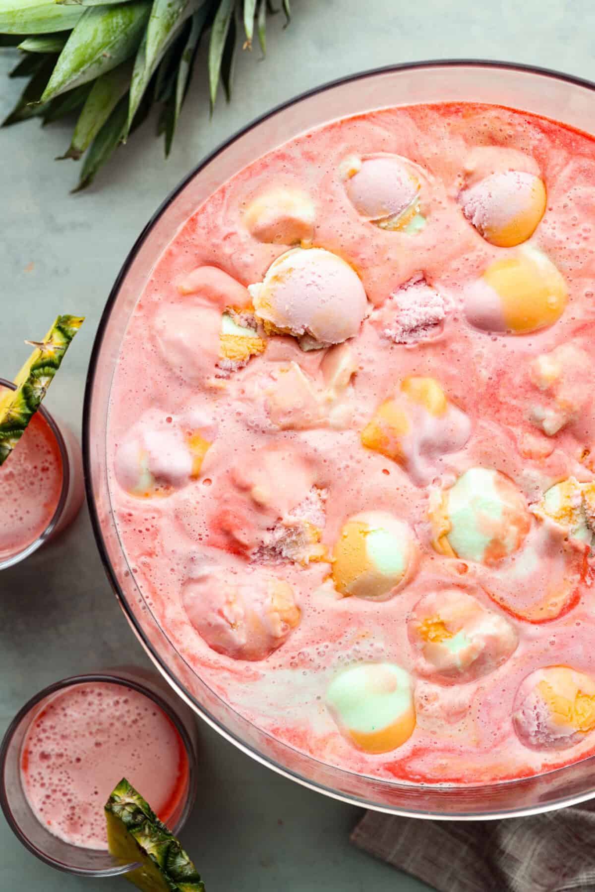 Overhead shot of a large punch bowl filled with sherbet punch next to smaller cups with the drink in them and a pineapple wedge on the rim of the glass.