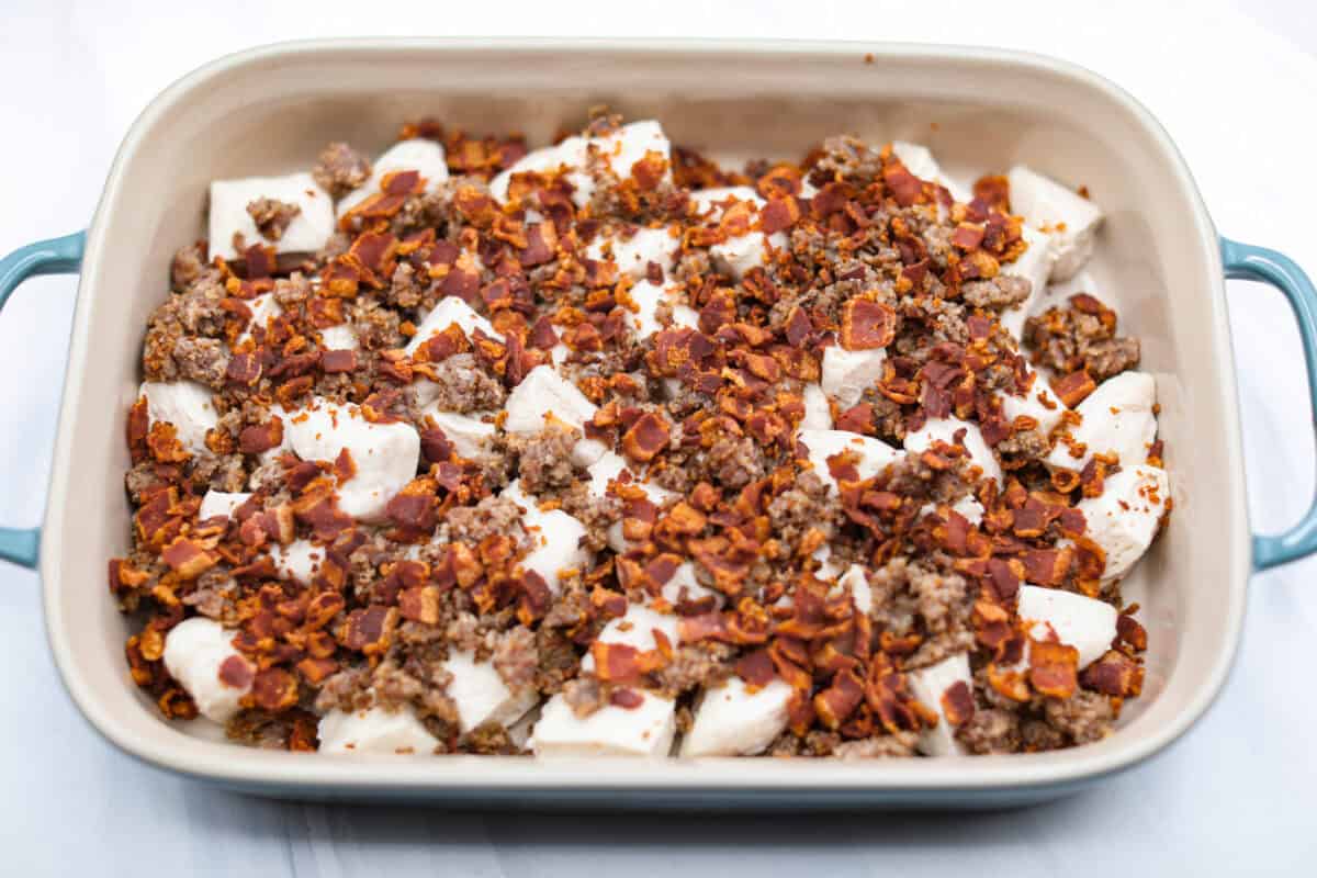 Overhead shot of bacon and sausage spread out over the biscuit pieces in the baking dish. 