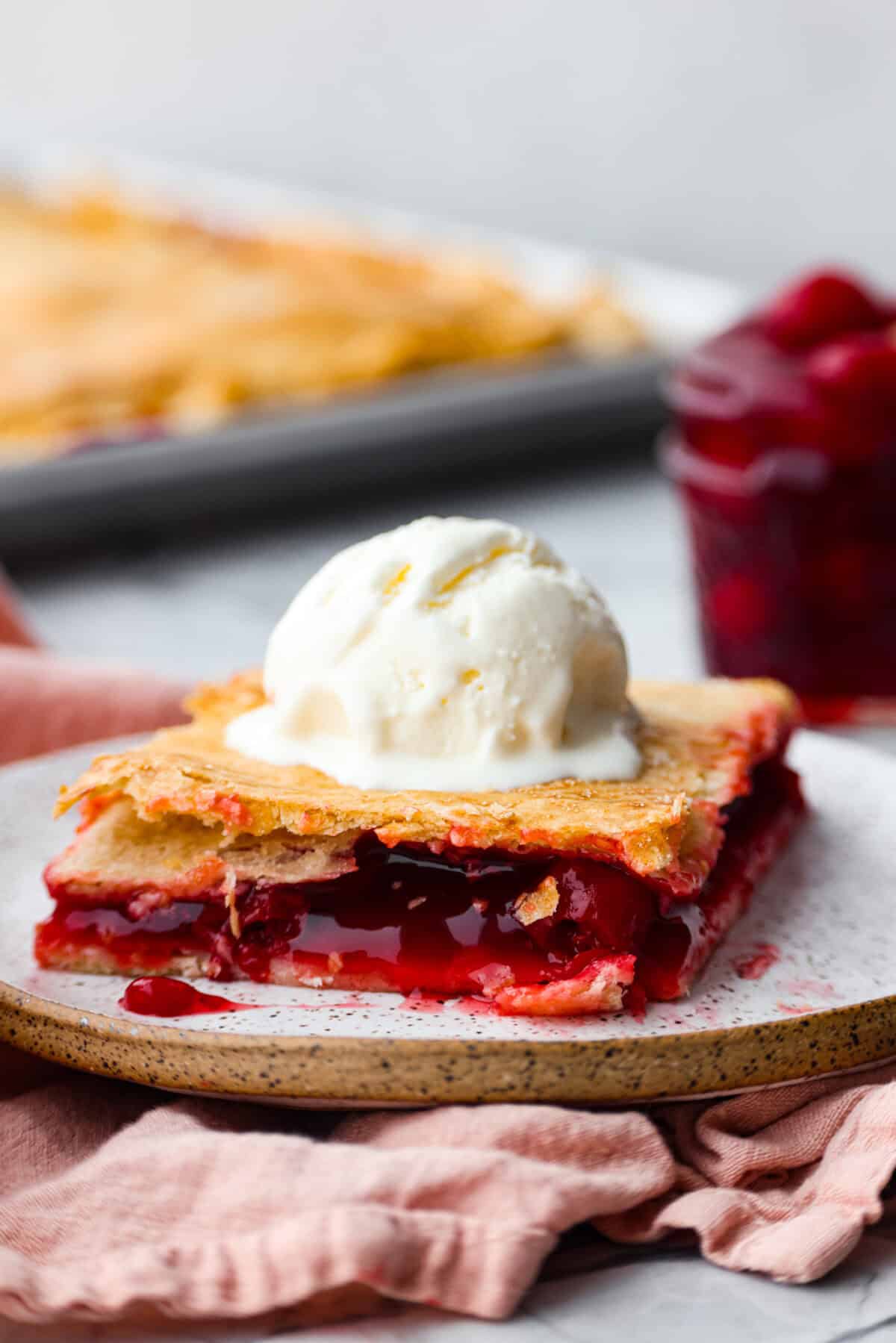 Side shot of a plated slice of cherry pie slab with a scoop of vanilla ice cream on top. 