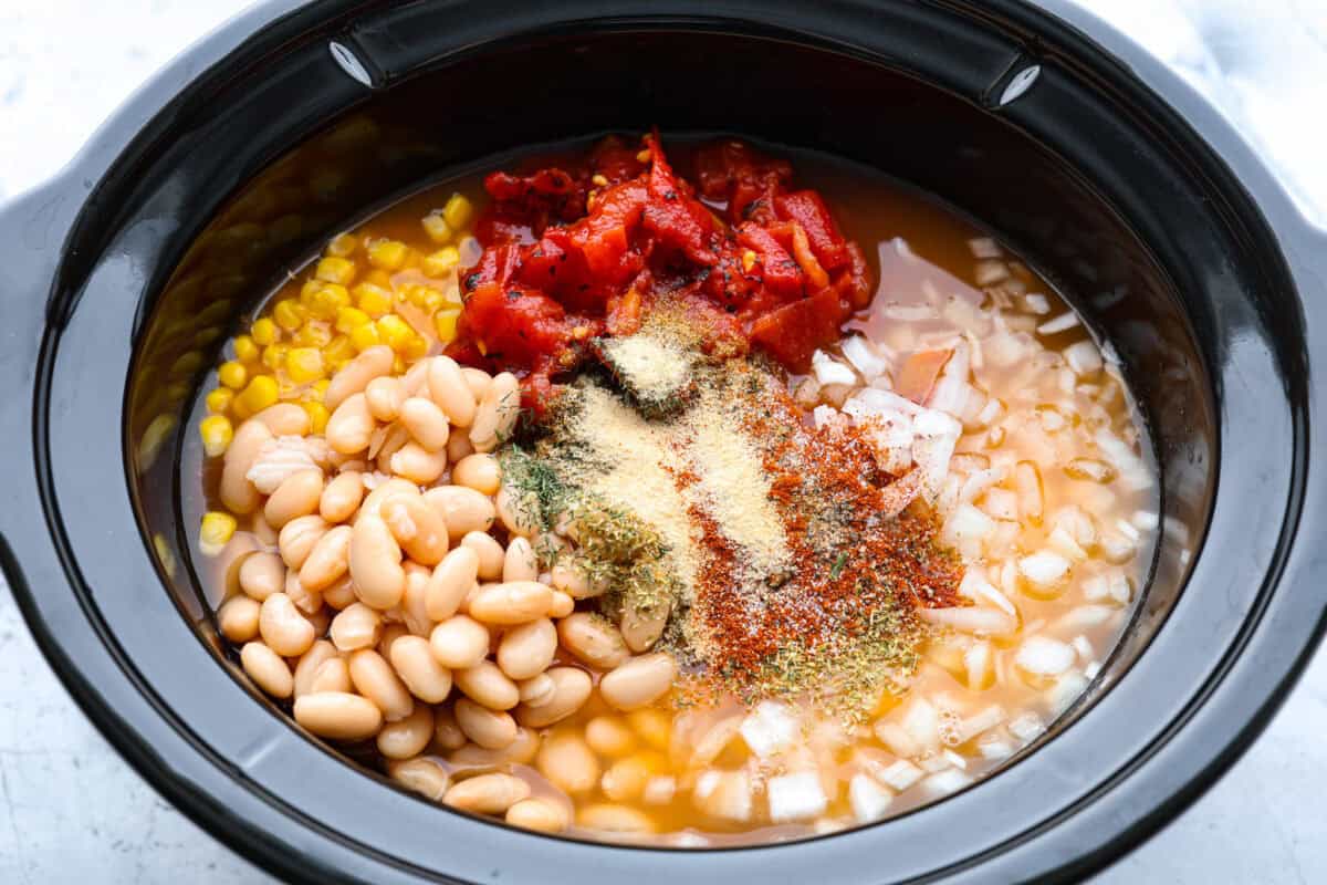 Overhead shot of the beans, corn, tomatoes, onion, buffalo sauce, chicken broth and seasonings in the crockpot.