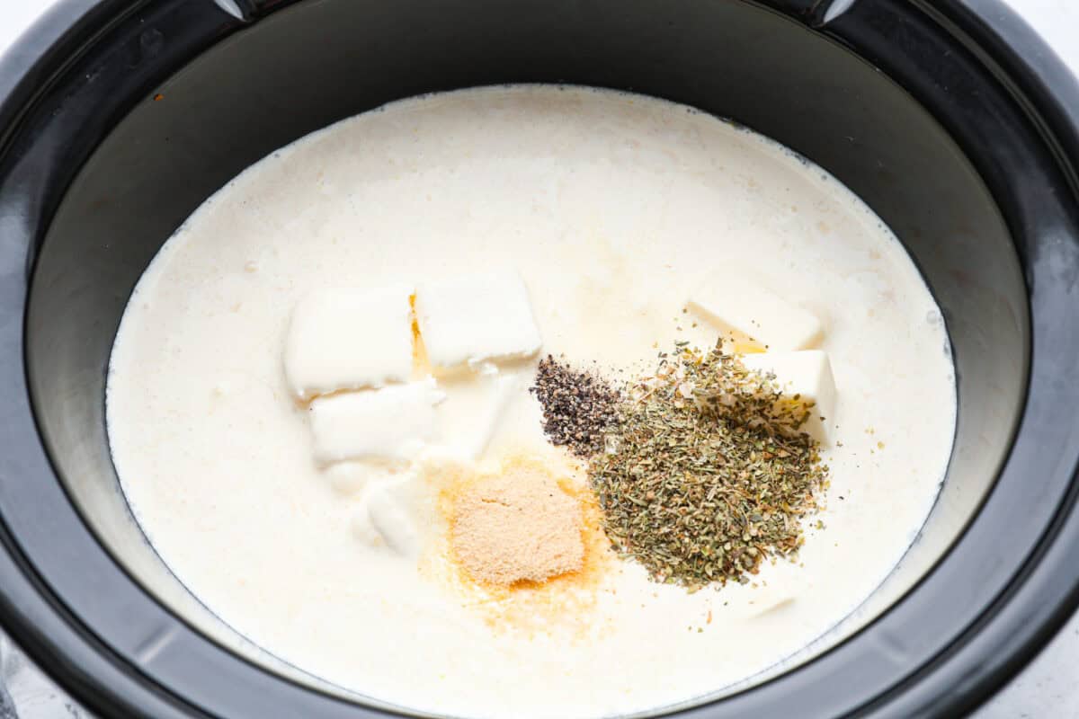 Overhead shot of chicken in the crockpot with seasonings, whipping cream, chicken broth, butter and cream cheese added on top. 