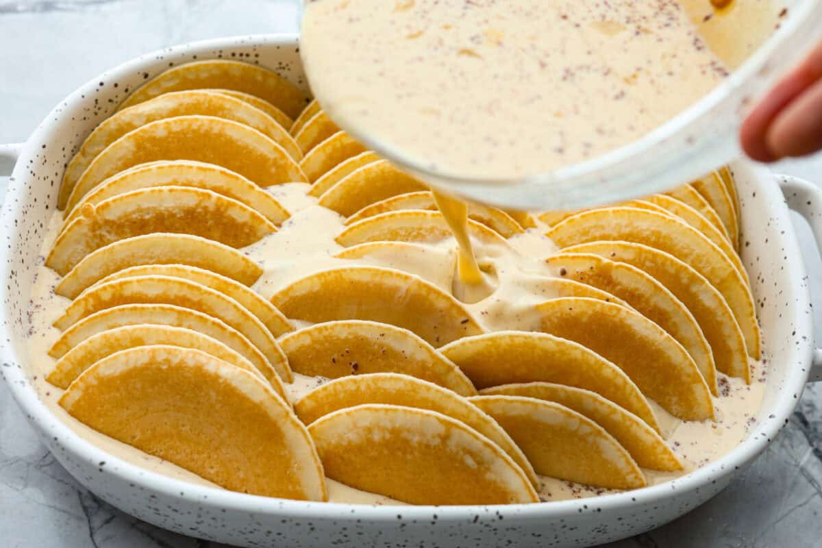 Angle shot of custard being poured over pancakes in baking dish. 