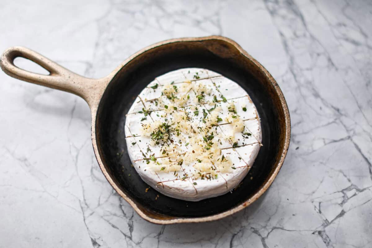 Overhead shot of the circle cheese with garlic and herbs sprinkled over the top of in a skillet. 