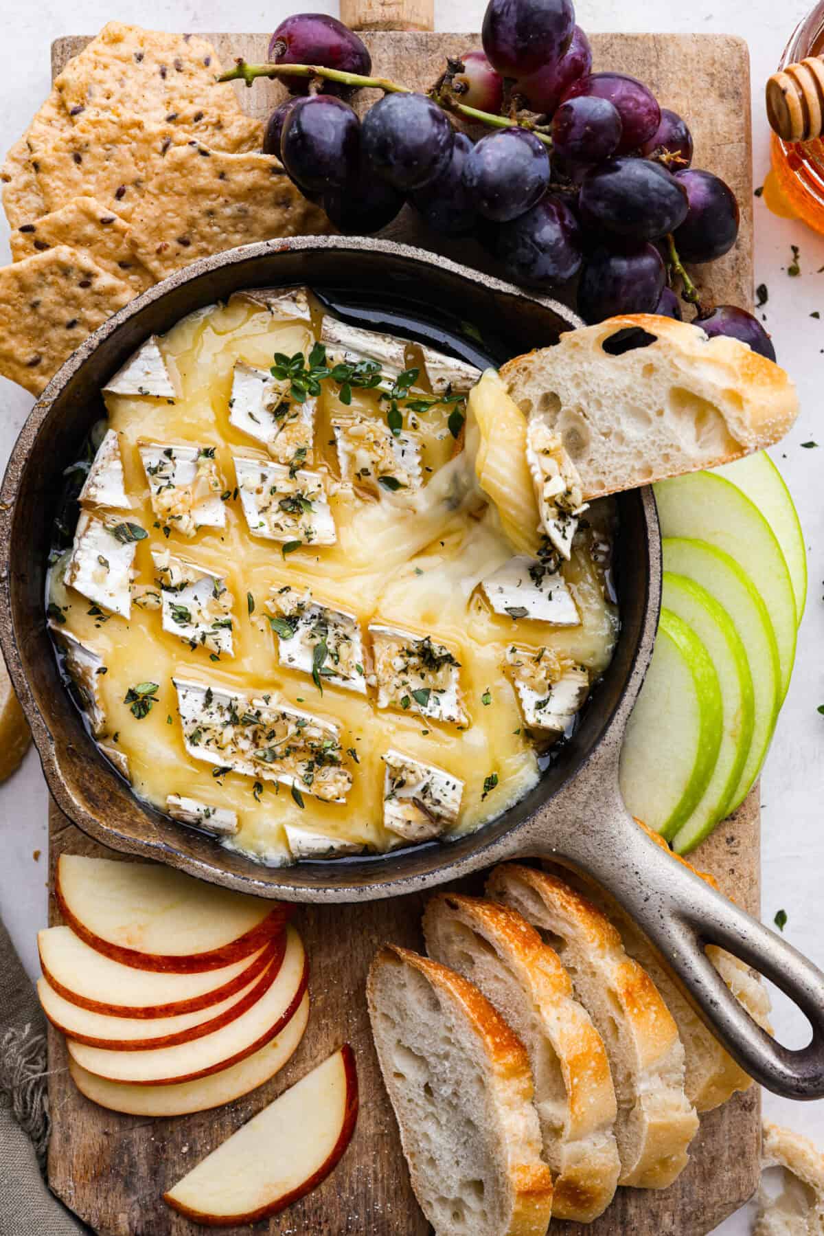 Overhead shot of baked brie on a cutting board with sliced apples, grapes, crackers and crostini. 