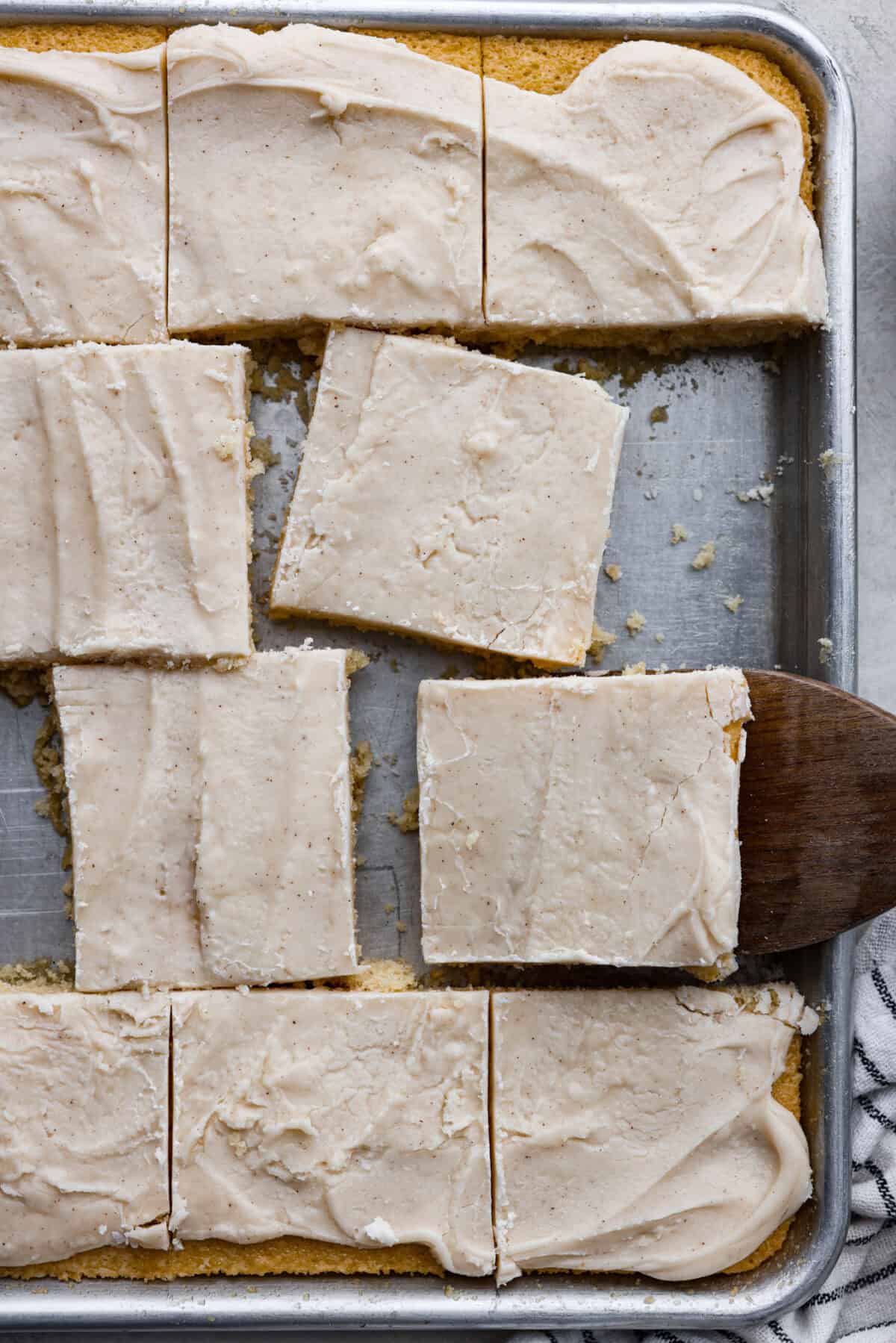 Overhead shot of browned butter sheet cake cut into squares in the baking sheet with someone taking one of the squares out with a wooden spatula.