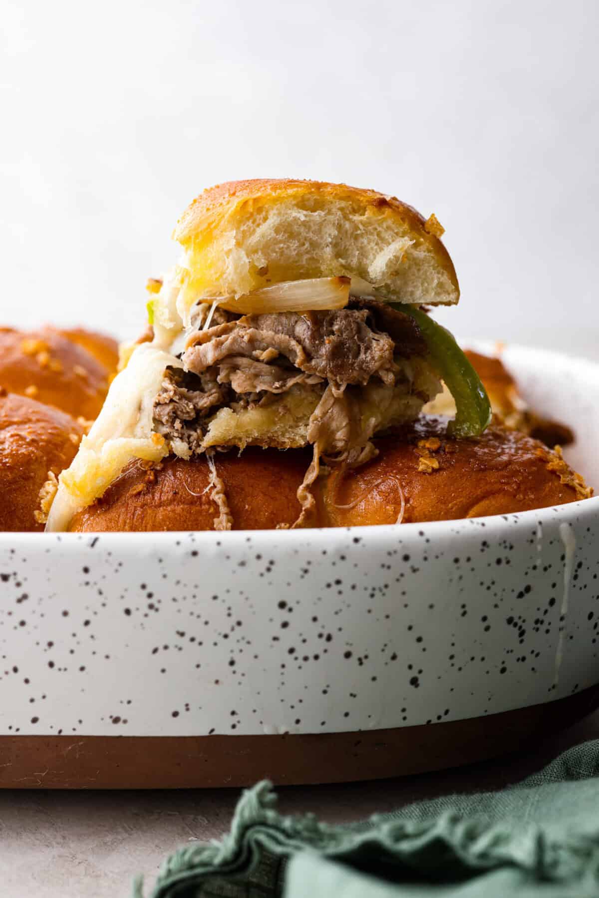 Side shot of a Philly cheesesteak slider on tope of the other sliders in a baking dish. 