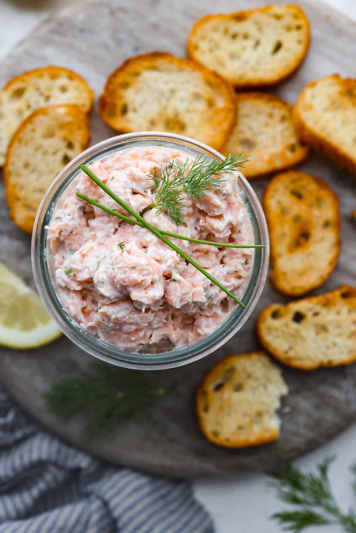 Overhead shot of salmon rillettes in a small bowl on a platter of crostini. 