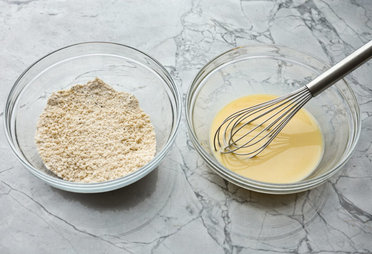 Angle shot of two glass bowl next to each other. One has breadcrumbs flour and seasoning, while the other has the milk and eggs whisked together. 