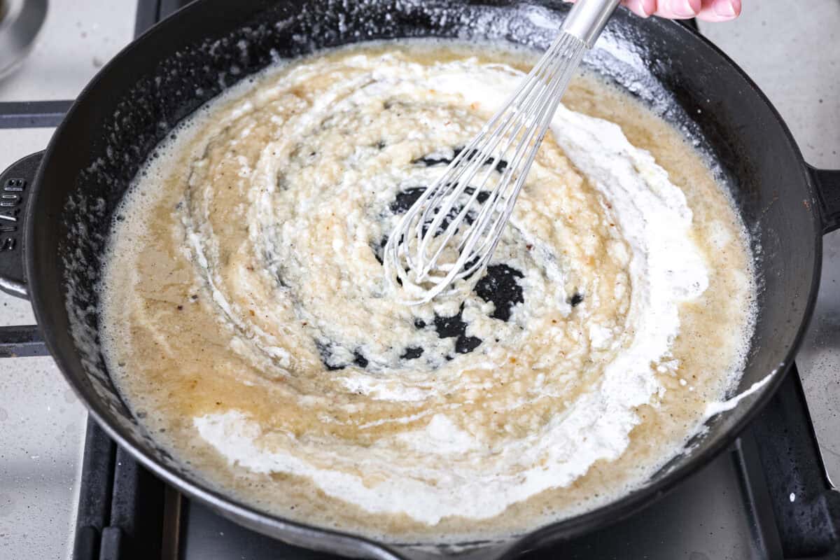 Angle shot of someone slowly adding in the chicken broth and cream mixture into the butter and flour mixture whisking continually. 