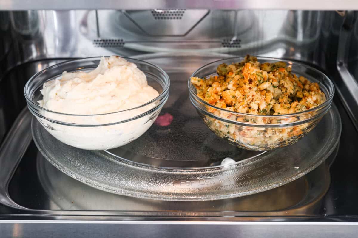 Side shot of a bowl of mashed potatoes next to a bowl of stuffing in a microwave. 