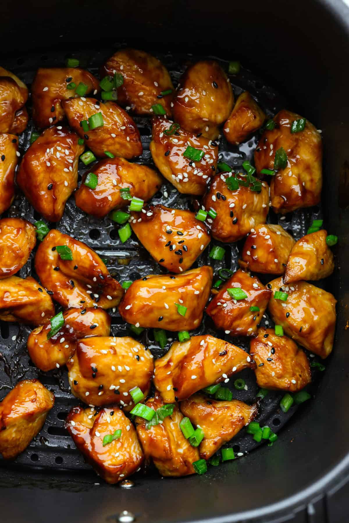 Close up shot of air fryer teriyaki chicken in the bottom of the air fryer basket with sesame seeds and green onions. 