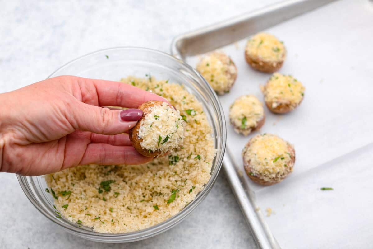 Overhead shot of someone holding a putting bread crumbs onto the cheese stuffed mushroom. 