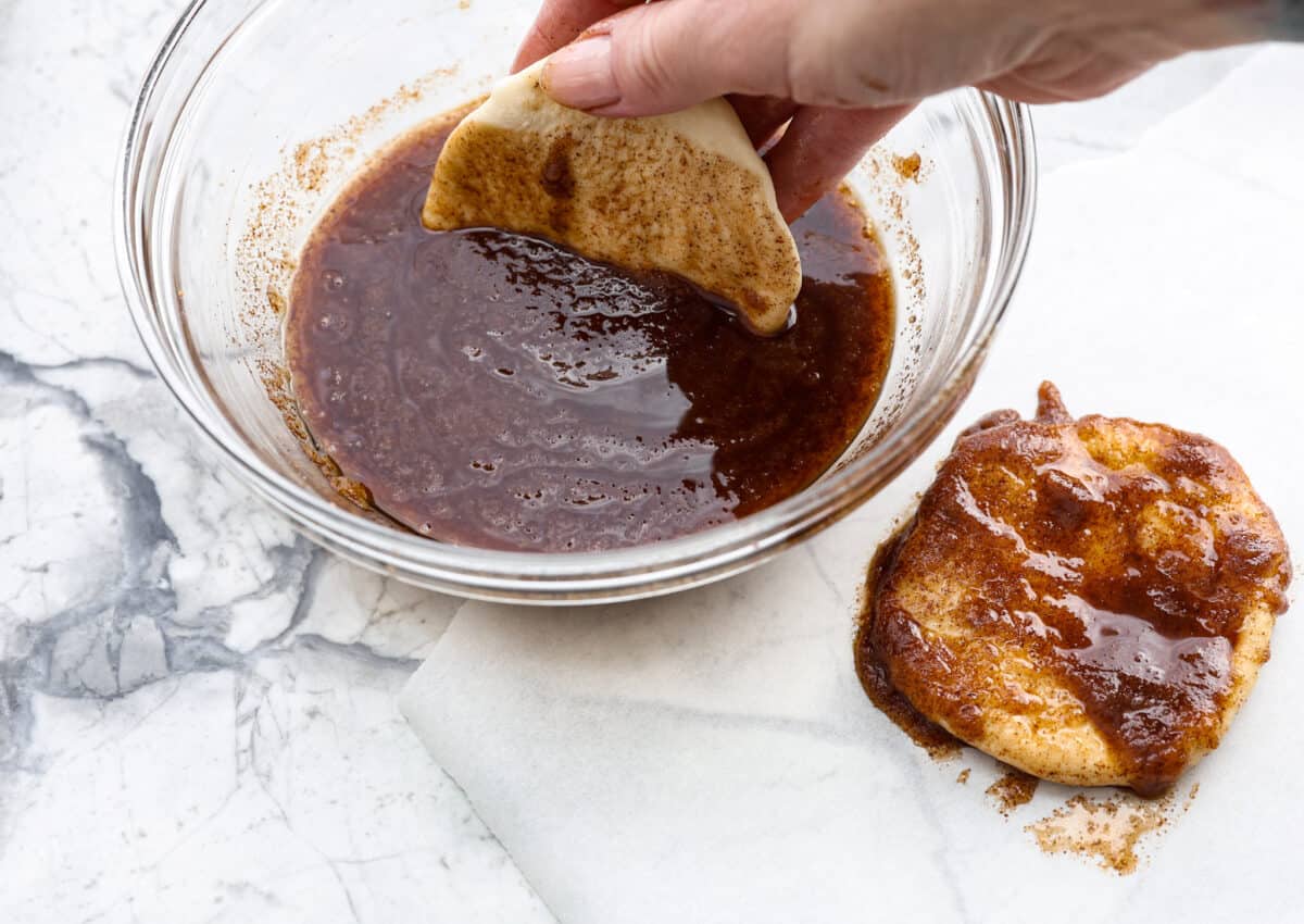 Angle shot of someone dipping the flattened dough into the butter, cinnamon and sugar mixture. 