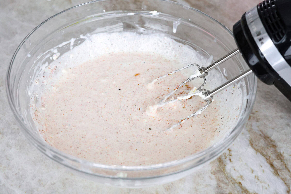 Angle shot of someone using a hand mixer to mix cream cheese, milk, garlic powder, paprika, salt and pepper. 