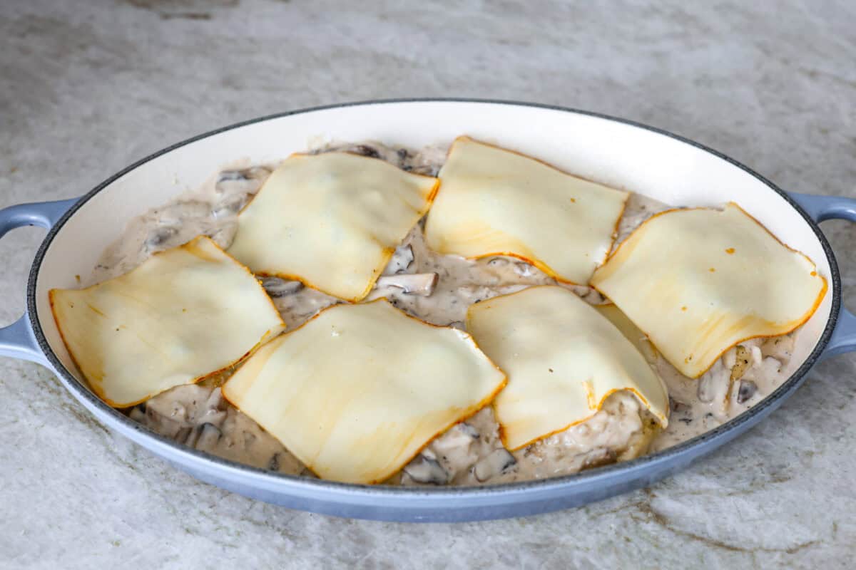 Angle shot of the chicken with the cream, broth and red wine vinegar in the sauce with mushrooms and broth in the baking dish covered with a slice of cheese on each chicken breast. 