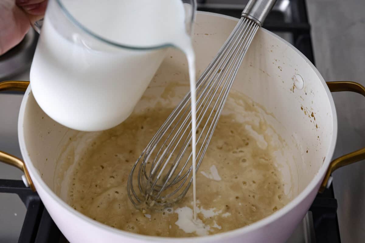 Angle shot of someone slowly pouring milk into the roux. 