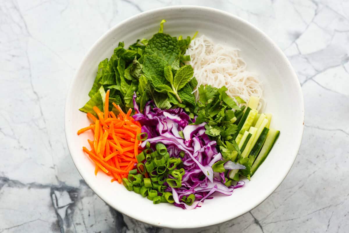 Overhead shot of a bowl with carrots, cabbage, cucumber, onion, cilantro lettuce, and mint on top of the noodles. 