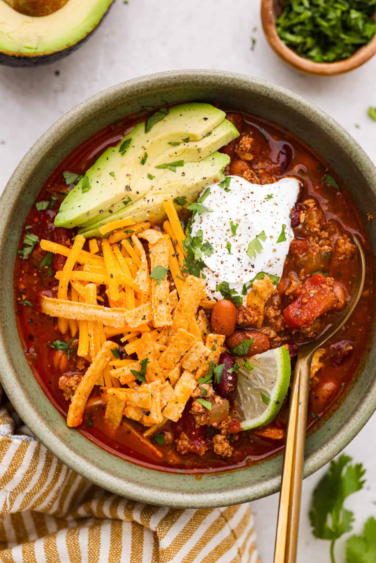 Overhead shot of a bowl of turkey chili garnished with sour cream, avocado, cheese, lime slice, and tortilla strips. 