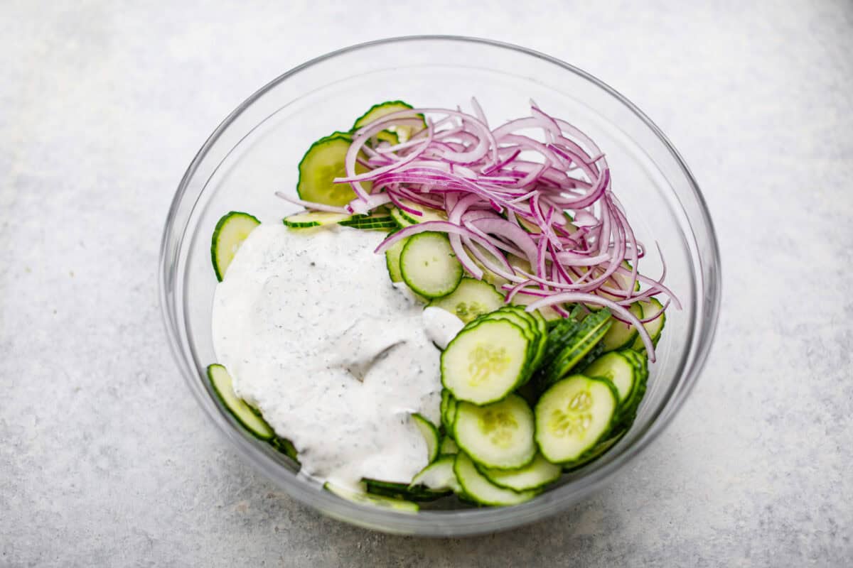 Overhead shot of sliced cucumbers, red onions and sauce in a bowl. 