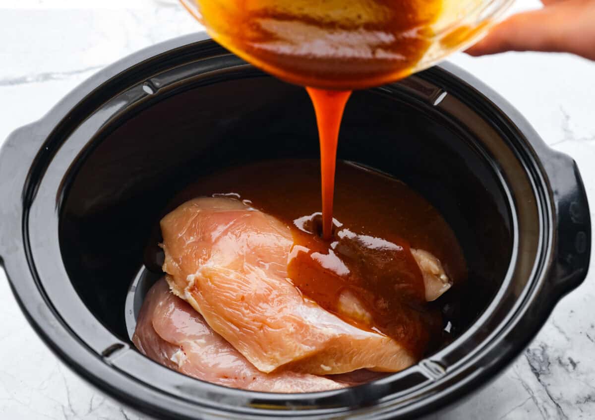 Angle shot of someone pouring the sauce over the chicken in the slow cooker. 