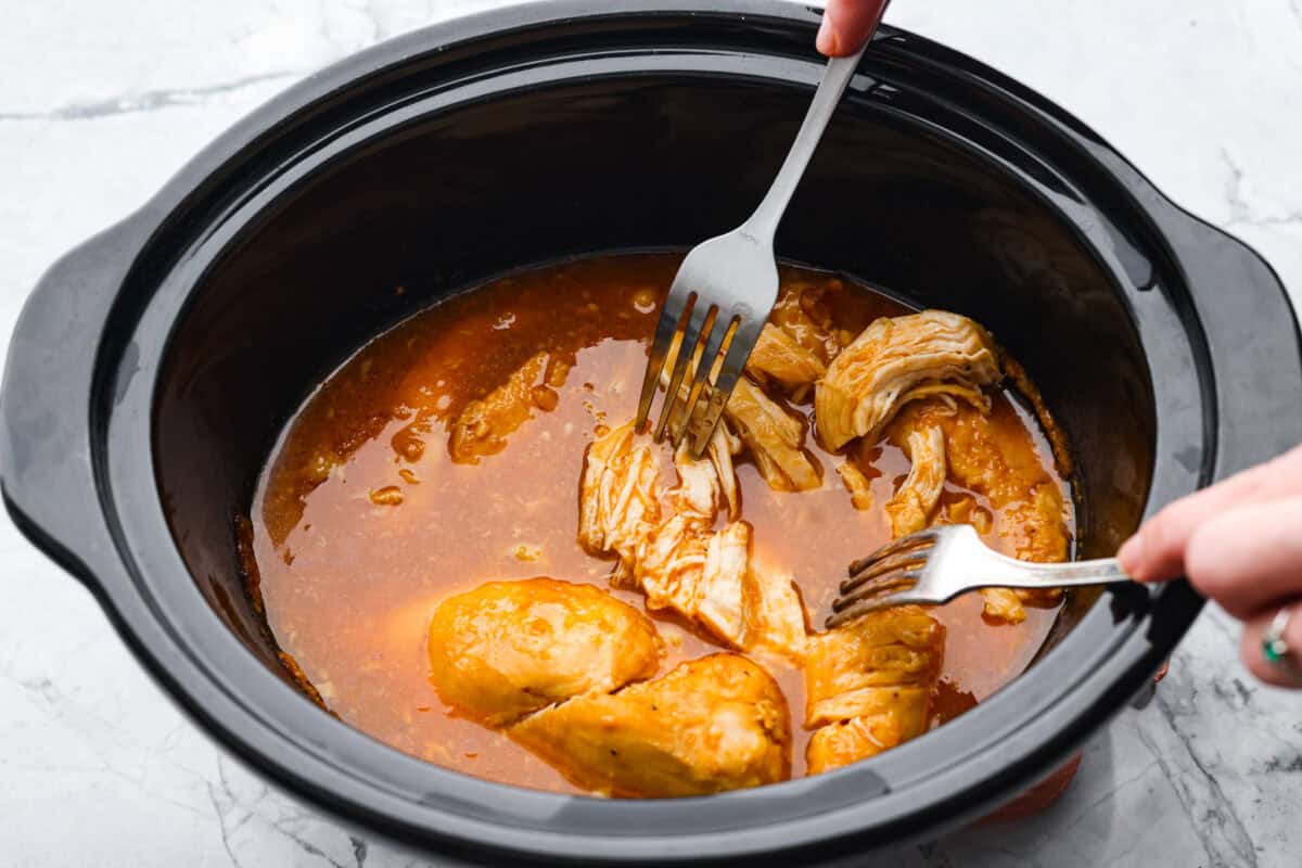 Overhead shot of someone shredding the chicken with two forks in the slow cooker. 